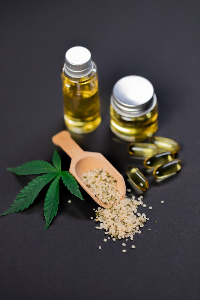 Flat lay of cannabis oil, hemp seeds, capsules, and leaf on dark background.